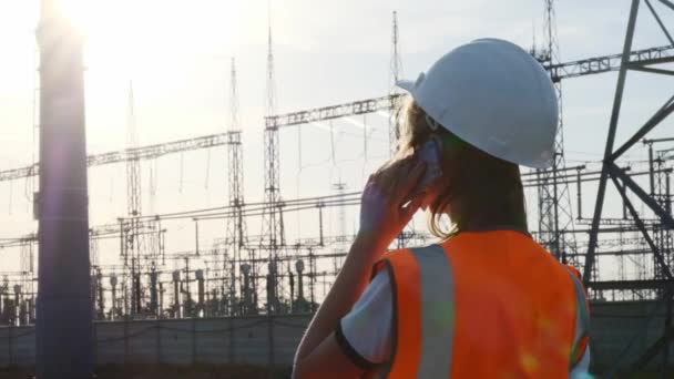 En flicka ingenjör i en vit hjälm och en orange skyddande väst inspekterar ett kraftverk med sladdar och elektricitet. Löser frågan om nya ägare. — Stockvideo
