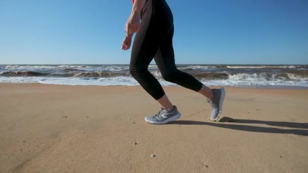 Une jeune fille court le long de la plage en tenue de sport rouge. Écoute de la musique sur écouteurs. Effectue un jogging le soir ou le matin au coucher du soleil ou à l'aube . — Video