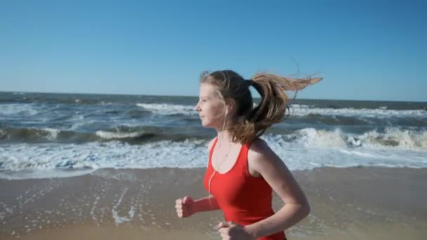 Retrato: uma jovem corre ao longo da praia em sportswear vermelho. Ouve música nos auscultadores. Executa uma noite ou uma corrida matinal ao pôr do sol ou ao amanhecer . — Vídeo de Stock