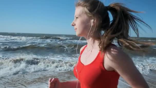 Porträtt: en ung flicka går längs stranden i röda sportkläder. Lyssnar på musik i hörlurar. Utför en kväll eller morgon jogga vid solnedgången eller dawn. — Stockvideo