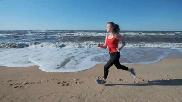 Πορτρέτο: ένα νεαρό κορίτσι που τρέχει κατά μήκος της παραλίας στο κόκκινο αθλητικών ειδών. Ακούει μουσική με ακουστικά. Εκτελεί ένα σκούντημα το βράδυ ή το πρωί στο ηλιοβασίλεμα ή την αυγή. — Αρχείο Βίντεο