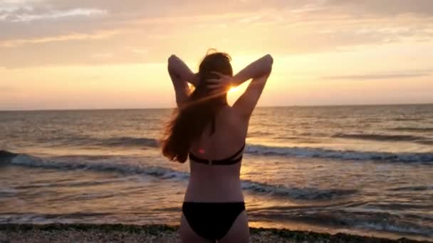 Menina na moda em um maiô preto entra no mar ao pôr do sol. Estou feliz com o mar e a praia. A experimentar a liberdade. Vai para a água. — Vídeo de Stock