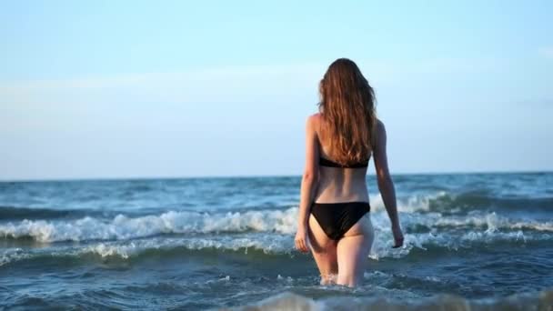 黒のファッショナブルな水着、セクシーな女の子が海に入ってくる。女の子は、足の上を歩く水に突入します。日没時、夜 — ストック動画