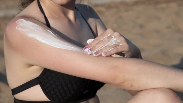 Сексуальна дівчина на пляжі в чорному купальнику змащена сонячним блоком або соляним кремом. Сидить на піску біля моря на пляжі. М'ясний крем на руці, плечі та ногах — стокове відео