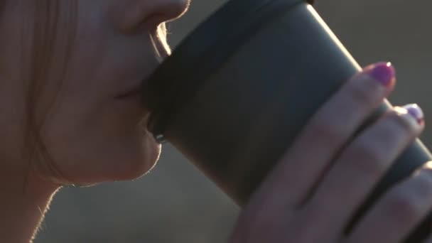 Menina sexy em um maiô preto beber café de um copo de papelão na praia. Noite ao pôr do sol ao lado do mar ou do oceano . — Vídeo de Stock