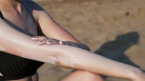 Menina sexy na praia em um maiô preto é manchado com protetor solar ou creme bronzeado do sol. Senta-se na areia junto ao mar na praia. Creme de esfregaço no braço, ombro e pernas — Vídeo de Stock