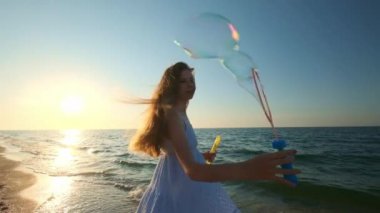 Kız plaj yavaş hareket baloncuklar üfleme. Genç bir kız akşamları, gün batımı sırasında sabun köpüğü darbeler. Plaj boyunca çalışır, sabun köpüğü güneş ışığında sinek..