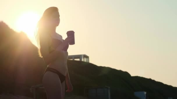 Sexy dziewczyna w czarny strój kąpielowy picia kawy z tektury szyby na plaży. Wieczorem zachód słońca obok morza lub oceanu. — Wideo stockowe