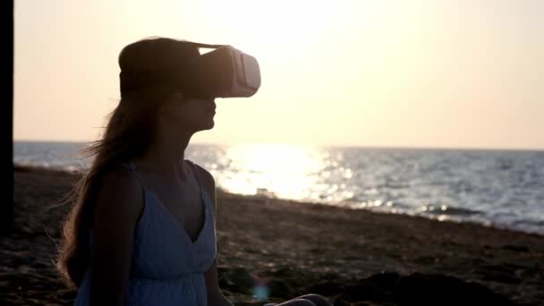 Młoda dziewczyna ubiera okulary wirtualnej rzeczywistości, Vr słuchawki. Rozlewa piasku się przez palce. Siedzi na piasku nad morzem o zachodzie słońca, na plaży. — Wideo stockowe