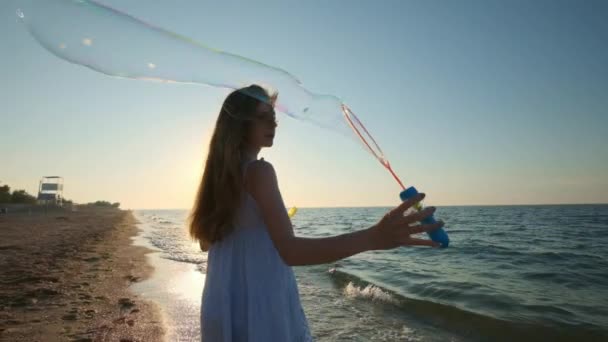 Fille soufflant des bulles sur la plage au ralenti. Une jeune fille souffle des bulles de savon le soir, au coucher du soleil. Cours le long de la plage, bulles de savon volent à la lumière du soleil . — Video