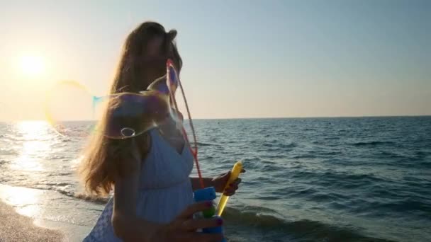 Meisje blazen van zeepbellen op strand in slow motion. Een jong meisje blaast zeepbellen in de avond, tijdens zonsondergang. Loopt langs het strand, zeepbellen vliegen in het zonlicht. — Stockvideo