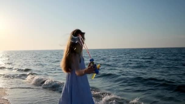 Dziewczyna dmuchanie baniek na plaży w zwolnionym tempie. Młoda dziewczyna wieje baniek mydlanych, wieczorem, podczas zachodu słońca. Biegnie wzdłuż plaży, bańki mydlane latać w słońcu. — Wideo stockowe