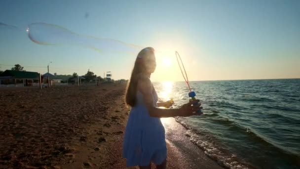 Dziewczyna dmuchanie baniek na plaży w zwolnionym tempie. Młoda dziewczyna wieje baniek mydlanych, wieczorem, podczas zachodu słońca. Biegnie wzdłuż plaży, bańki mydlane latać w słońcu. — Wideo stockowe