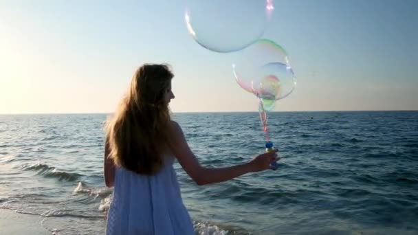 Flicka blåser bubblor på stranden i slow motion. En ung flicka blåser såpbubblor på kvällen under solnedgången. Löper längs stranden, såpbubblor flyger i solljus. — Stockvideo