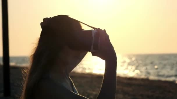 Молодая женщина использует очки виртуальной реальности на пляже. Сексуальная девушка получает опыт использования гарнитуры летом на морском фоне . — стоковое видео