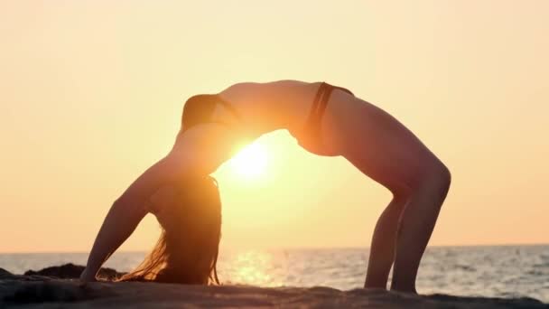 Młoda kobieta w garniturze ćwiczy jogę na plaży nad morzem o niesamowitym wschodzie słońca. Fitness, sport, joga i koncepcja zdrowego stylu życia. Dziewczyna robi most, zwolniony ruch. — Wideo stockowe