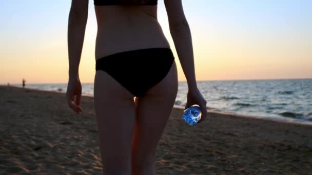 Ein sportliches Mädchen mit einer Flasche auf dem Weg zum Meer. sexy Mädchen, das in einem schwarzen Badeanzug am Strand entlang läuft. in ihren Händen hält eine Flasche sauberes Wasser. — Stockvideo