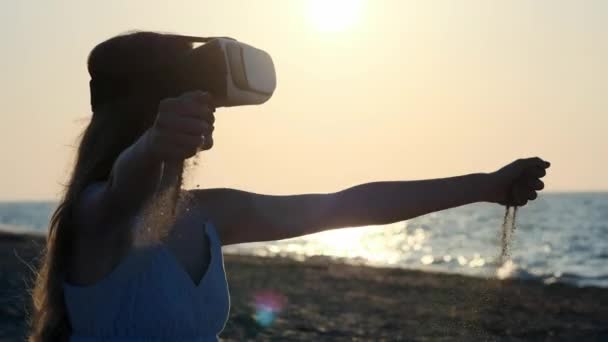 Ragazza veste occhiali di realtà virtuale, VR-headset. Versa la sabbia tra le dita. Seduto sulla sabbia in riva al mare al tramonto, sulla spiaggia . — Video Stock