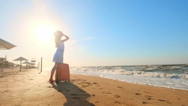 Молода жінка з валізою сидить на пляжі. молода дівчина ходить уздовж пляжу з колісною валізою. Дівчина шукає себе і пригоди . — стокове відео