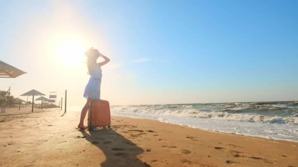 Wanita muda dengan koper duduk di pantai. seorang gadis muda berjalan di sepanjang pantai dengan koper roda. Shags sepanjang pasir di sepanjang laut. Gadis itu mencari dirinya sendiri dan petualangan . — Stok Video