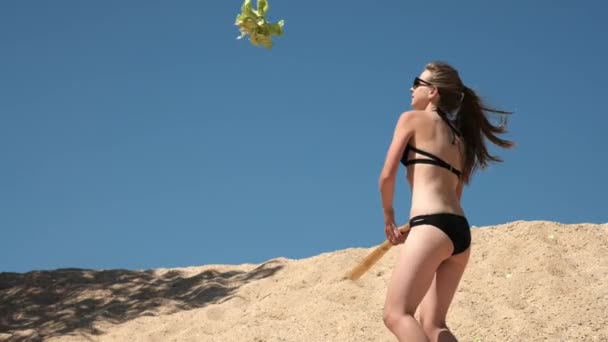 Медленное движение: Сексуальная молодая девушка в черном купальнике и очках, уничтожает фрукты и овощи бейсбольной битой. Он расположен на песчаном пляже у моря в летнее время. Разбивает капусту . — стоковое видео