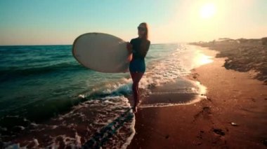 Ağır çekim: surfboard holding sahilde yürüyüş güzel ince kız. Akşam gün batımı sırasında çalışır. Dalış elbisesi, koruyucu su geçirmez elbise giyer yüzme bir uygun