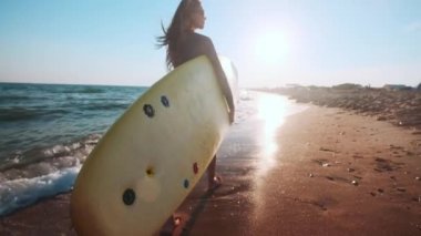 Ağır çekim: surfboard holding sahilde yürüyüş güzel ince kız. Akşam gün batımı sırasında çalışır. Dalış elbisesi, koruyucu su geçirmez elbise giyer yüzme bir uygun
