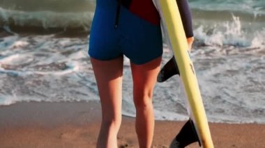 Ağır çekim: Deniz sahil boyunca ince kız profesyonel sörfçü gelir. Sarı sörf tahtaları elinde bir wetsuit giyinmiş.