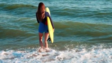 Ağır çekim: Deniz sahil boyunca ince kız profesyonel sörfçü gelir. Sarı sörf tahtaları elinde bir wetsuit giyinmiş.