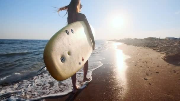 LOW MOTION: Menina magro bonita andando na praia, segurando prancha de surf. É executado à noite durante o pôr do sol. Usa um fato de banho, um fato impermeável de protecção — Vídeo de Stock