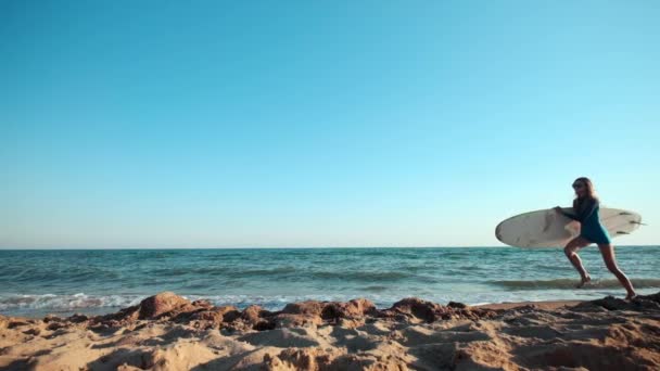 Zwolnionym tempie: Piękna szczupła dziewczyna spaceru na plaży, trzymając deskę surfingową. Działa w godzinach wieczornych podczas zachodu słońca. Nosi widoki dostosowane kombinezon, strój ochronny wodoodporny — Wideo stockowe