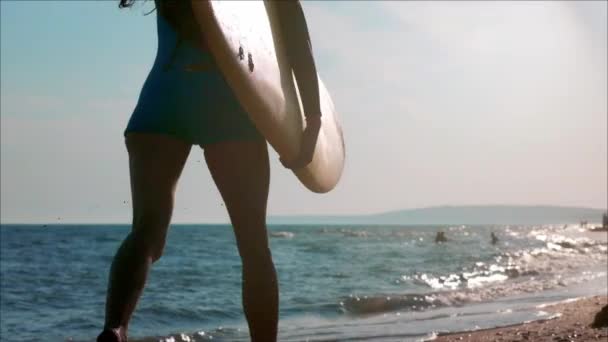 Гарна струнка дівчина, що йде на пляжі, тримає дошку для серфінгу. Біжить увечері під час заходу сонця. Носить купальник, захисний водонепроникний костюм — стокове відео