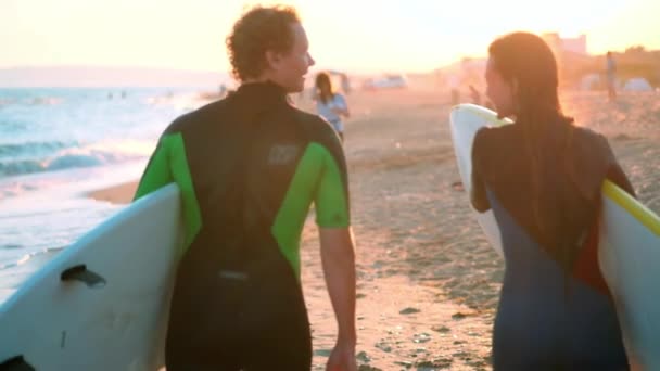 Slow Motion: Een jong stel van jongen en meisje surfers zijn wandeling langs het strand in wetsuits. In de handen van holding surfplanken. De golven en de ondergaande zon kijken. — Stockvideo