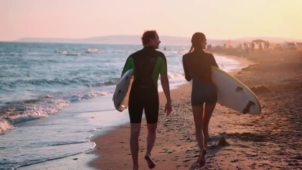 Медленное движение: молодая пара парней и девочек-серферов прогуливаются по пляжу в гидрокостюмах. В руках у досок для сёрфинга. Посмотри на волны и закат . — стоковое видео