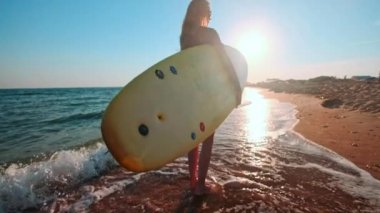 Ağır çekim: surfboard holding sahilde yürüyüş güzel ince kız. Akşam gün batımı sırasında çalışır. Dalış elbisesi, koruyucu su geçirmez elbise giyer yüzme bir uygun