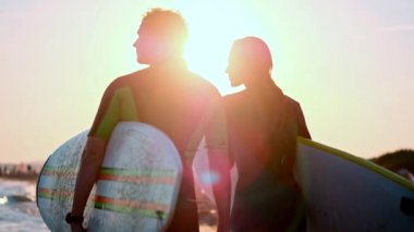 Ağır çekim: Genç bir çift adam ve kız sörfçü wetsuits plaj boyunca yürüme. Sörf tahtaları tutarak ellerinde. Dalgalar ve gün batımı bak.