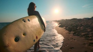 Ağır çekim: surfboard holding sahilde yürüyüş güzel ince kız. Akşam gün batımı sırasında çalışır. Dalış elbisesi, koruyucu su geçirmez elbise giyer yüzme bir uygun