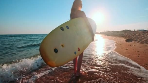 Slowmotion: Vacker smal tjej promenader på stranden, hålla surfbräda. Körs på kvällen under solnedgången. Bär en simning passar våtdräkt, vattentät skyddsdräkt — Stockvideo