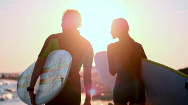 Slow Motion: Een jong stel van jongen en meisje surfers zijn wandeling langs het strand in wetsuits. In de handen van holding surfplanken. De golven en de ondergaande zon kijken. — Stockvideo