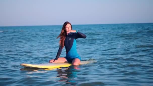 MOCIÓN LENTA: Joven chica delgada de surf mirando el atardecer. Flota en una tabla amarilla en el mar abierto, espera una ola y mira al horizonte. Lleva un traje de neopreno profesional . — Vídeos de Stock