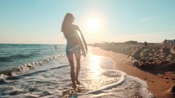 Красива струнка дівчина, що йде на пляжі, тримає дошку для серфінгу. Біжить увечері під час заходу сонця. Носить купальник, захисний водонепроникний костюм — стокове відео