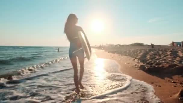 Beautiful slim girl walking on the beach, holding surfboard. Runs in the evening during sunset. Wears a swimming suit wetsuit, protective waterproof suit — Stock Video