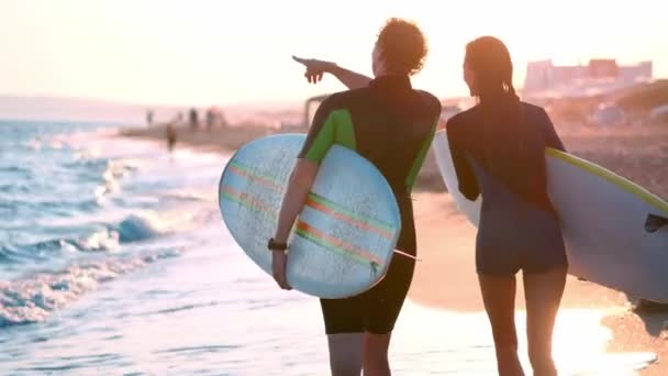Um jovem casal de surfistas e surfistas anda pela praia de fato de mergulho. Nas mãos de segurar pranchas de surf. Olha para as ondas e o pôr-do-sol . — Vídeo de Stock