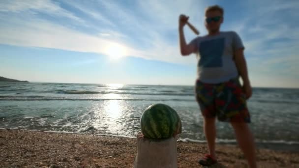 Cámara lenta: un joven sexy golpea a un bate de béisbol con verduras y una fruta de sandía. Se encuentra en la playa, playa de arena — Vídeo de stock