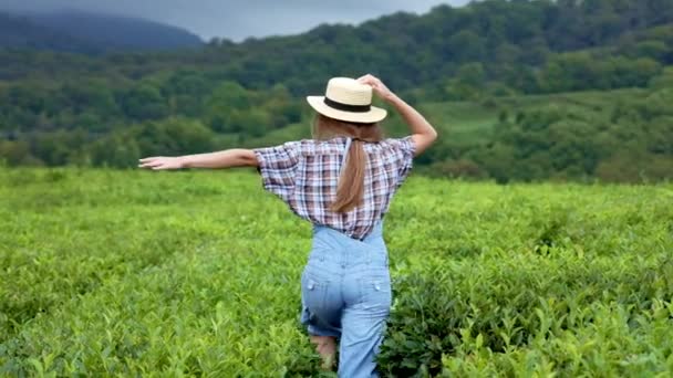 Une jeune belle paysanne en costume de jeans et un chapeau de paille court le long d'une plantation de thé ou de café. Vivre le bonheur de voyager, l'unité avec la nature . — Video