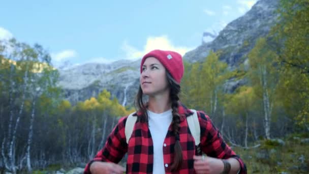 Chica hipster joven activa viaja a través del bosque, va a lo largo del camino entre las montañas. Lleva un sombrero rojo y una camisa roja a cuadros. Mochila de senderismo en los hombros . — Vídeos de Stock