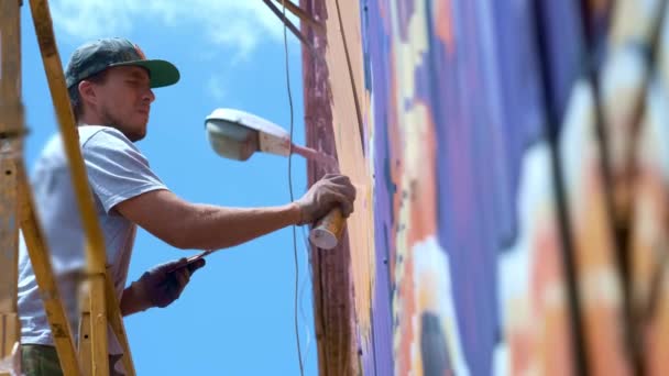 Vista bassa: Un giovane artista di sesso maschile disegna una lattina di pittura a graffiti su una parete. Dovrebbe essere su una torre di costruzione. Un uomo impegnato nell'arte . — Video Stock