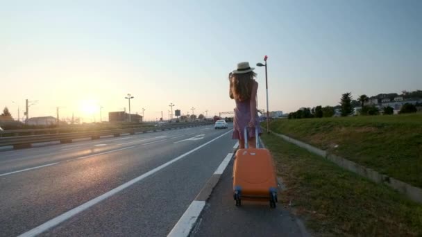 Dziewczyna idzie na drodze z walizką. W pobliżu autostrady i wiele samochodów.. — Wideo stockowe
