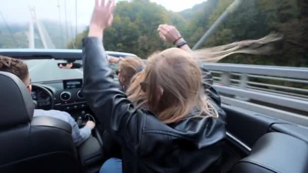 Dos hermosas jóvenes viajan en un cabriolet rojo entre las montañas. Camino en la carretera. Vestido con chaquetas de cuero negro. Cabello revoloteando en el viento. Disfruta de viajes y libertad . — Vídeo de stock