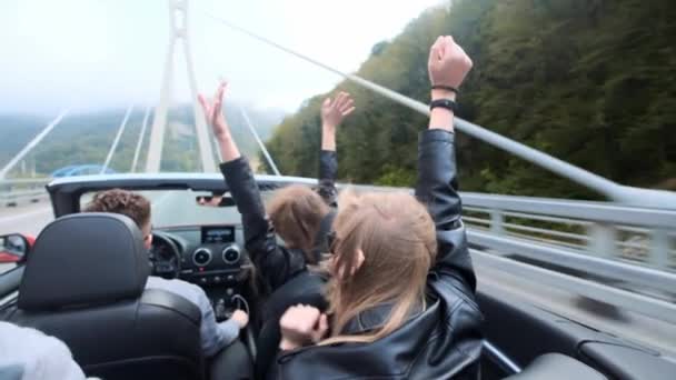 Две красивые молодые девушки ездят в красной кабриолете среди гор. Дорога по шоссе. Одетые в черные кожаные куртки. Волосы трепещут на ветру. Наслаждайтесь путешествиями и свободой . — стоковое видео